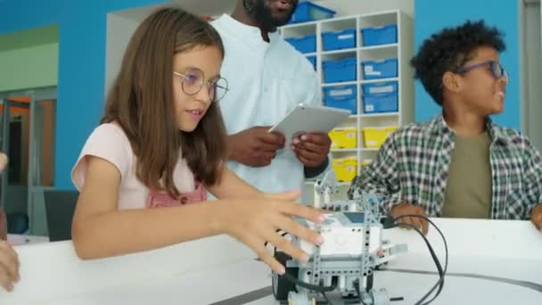 Μέτριο πλάνο της ομάδας των νέων διαφορετικών παιδιών και μαύρο αρσενικό δάσκαλο παίζει με προγραμματιζόμενα ηλεκτρικά ρομπότ στο μετά το σχολείο tech club, και εκστατικό αγόρι άλμα με χαρά στην ολοκλήρωση της εργασίας - Πλάνα, βίντεο