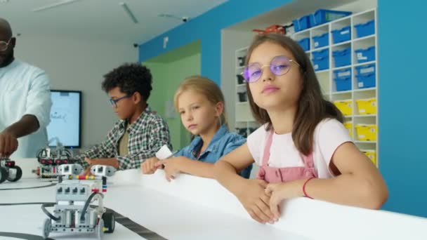 Μεσαία φωτογραφία πορτραίτου ενός σπασίκλα προ εφήβου καυκάσιου κοριτσιού με γυαλιά που στέκεται στο τραπέζι στο νεανικό κλαμπ ρομποτικής και χαμογελά στην κάμερα, ενώ συμμαθητές και δάσκαλοι χειρίζονται ηλεκτρικά μοντέλα ρομπότ - Πλάνα, βίντεο