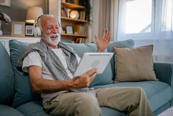 Een oudere man oude blanke man gepensioneerde zitten thuis op de slaapbank gebruik digitale tablet om film te kijken of hebben een video gesprek online met koptelefoon op zijn hoofd grijs haar en baard - Foto, afbeelding