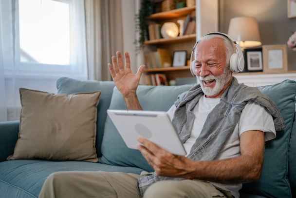Jeden starszy mężczyzna stary biały mężczyzna emeryt siedzieć w domu na kanapie za pomocą tabletu cyfrowego do oglądania filmów lub mieć wideo połączenie online ze słuchawkami na głowie siwe włosy i broda - Zdjęcie, obraz