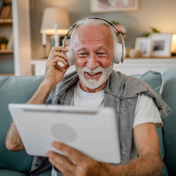 Een oudere man oude blanke man gepensioneerde zitten thuis op de slaapbank gebruik digitale tablet om film te kijken of hebben een video gesprek online met koptelefoon op zijn hoofd grijs haar en baard - Foto, afbeelding