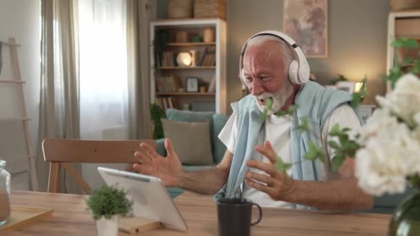 Een oudere man oude blanke man gepensioneerde zitten thuis op de slaapbank gebruik digitale tablet om film te kijken of hebben een video gesprek online met koptelefoon op zijn hoofd grijs haar en baard - Video