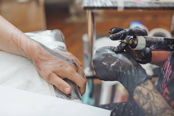 przycięty strzał procesu tatuażu na rękę w salonie. Tatuażysta wprowadza atrament do skóry za pomocą igły z tatuaża.Profesjonalny tatuażysta pracujący w studio. Skupienie selektywne - Zdjęcie, obraz
