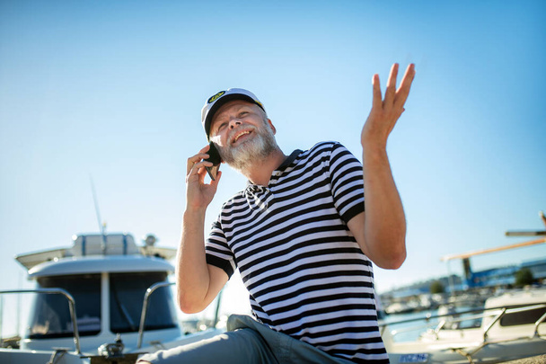 Homme mûr debout près de la mer vêtu d'une chemise et d'un chapeau de marin en utilisant un téléphone intelligent - Photo, image