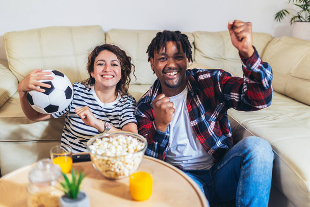 Afroamerikaner und Latina-Frau sehen sich ein Fußballspiel im Fernsehen bequem von zu Hause aus an. - Foto, Bild