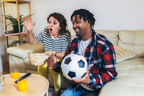 Афроамериканский муж и латиноамериканская жена смотрят футбол по телевизору из комфорта своего дома. - Фото, изображение