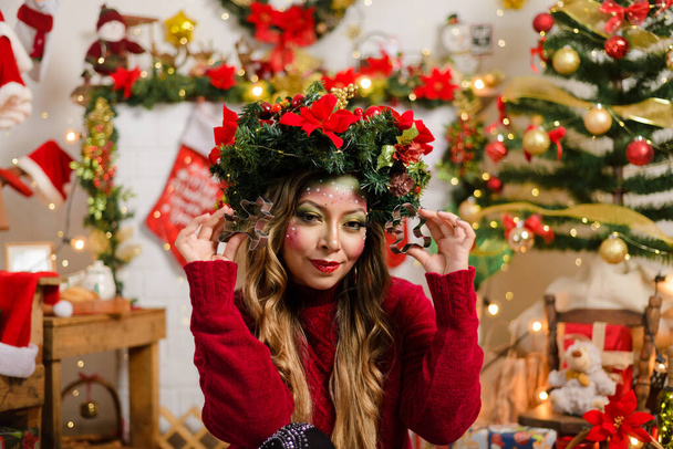 Beautiful woman with Christmas makeup wearing Christmas wreath and red sweater sitting by fireplace in living room decorated for Christmas, holding cookie cutter. - Photo, Image