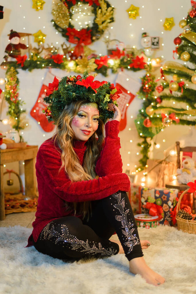 Beautiful woman with Christmas makeup wearing Christmas wreath and red sweater seated by fireplace in living room decorated for Christmas. - Photo, Image