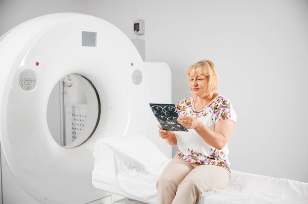 Medical computed tomography or MRI scanner. Patient making MRI, sitting on couch. Blonde woman holding MRI result, scan, x-ray, looking, examining, thinking. Concept of medicine and healthcare. - Photo, Image
