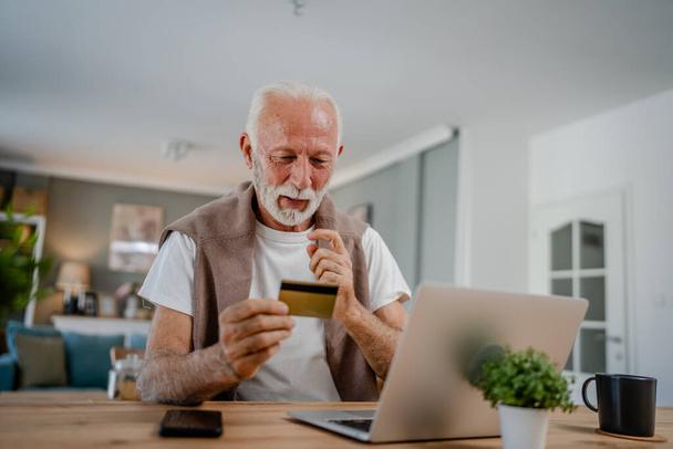 Um homem do sexo masculino caucasiano sênior avô sentar em casa usar cartão de crédito e computador portátil compras on-line usar internet para compra on-line ativo moderno sênior - Foto, Imagem