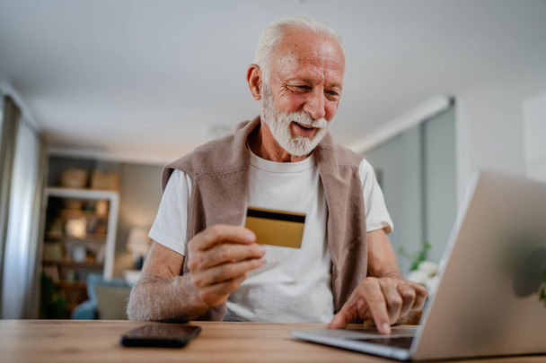 jeden człowiek starszy kaukaski mężczyzna dziadek siedzieć w domu korzystać z karty kredytowej i laptopa zakupy online korzystać z Internetu dla zakupu online aktywny nowoczesny senior - Zdjęcie, obraz