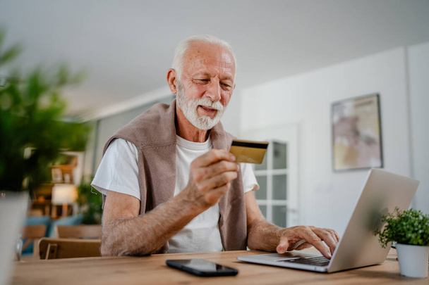 Üst düzey bir beyaz erkek büyükbaba evde oturuyor. İnternette kredi kartı ve dizüstü bilgisayar alışverişi yapıyor. İnternetten modern son sınıf öğrencilerini satın almak için internet kullanıyor. - Fotoğraf, Görsel