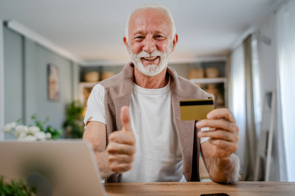 Um homem do sexo masculino caucasiano sênior avô sentar em casa usar cartão de crédito e computador portátil compras on-line usar internet para compra on-line ativo moderno sênior - Foto, Imagem
