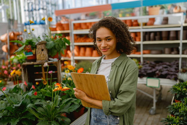 Smiling woman florist with clipboard conducts inventory in flower garden center  - Photo, Image