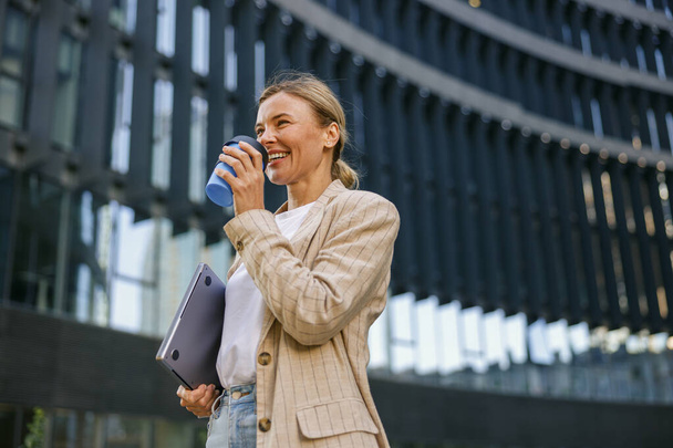 Gerente feminina positiva bebendo café durante o intervalo perto do prédio de escritórios. Foto de alta qualidade - Foto, Imagem
