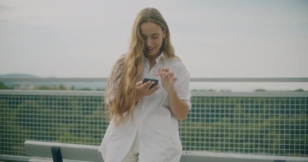 Lassú mozgás lövés vonzó szőke nő élvezi a zenét keresztül okos telefon, miközben támaszkodik padon - Felvétel, videó