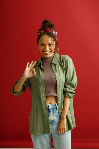 Femme montrant signe Ok avec le sourire tout en se tenant sur fond rouge studio. Photo de haute qualité - Photo, image