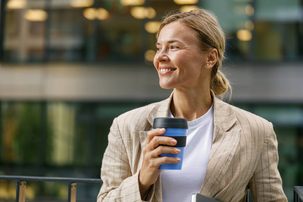 Gerente feminina positiva bebendo café durante o intervalo perto do prédio de escritórios. Foto de alta qualidade - Foto, Imagem
