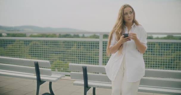 Медленный снимок задумчивой молодой женщины, держащей смартфон, опираясь на скамейку - Кадры, видео