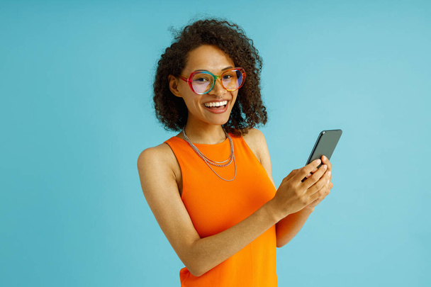 Красива жінка в окулярах використовує мобільний телефон, стоячи на синьому фоні студії  - Фото, зображення