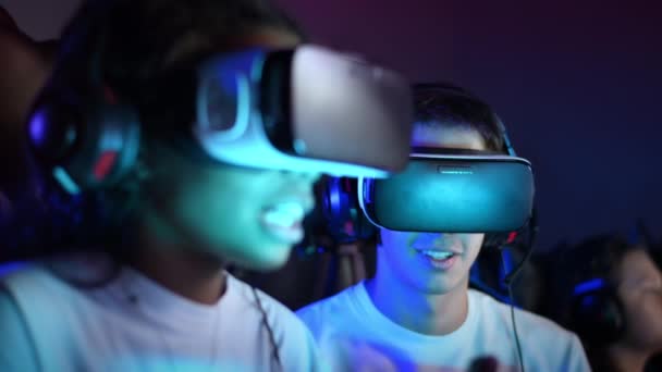 Adolescentes blancos y negros con auriculares VR jugando videojuegos en un club de videojuegos con iluminación usando un mando, hablando en chat de voz. Movimiento lento - Imágenes, Vídeo