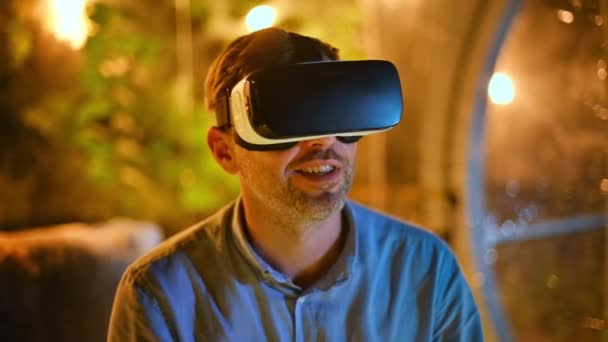 Hombre hablador en gafas VR sentado dentro de una tienda de glamping con iluminación por la noche - Metraje, vídeo