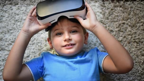 Bionda piccolo ragazzo sdraiato sul tappeto mostrando come e indossando VR auricolare - Filmati, video