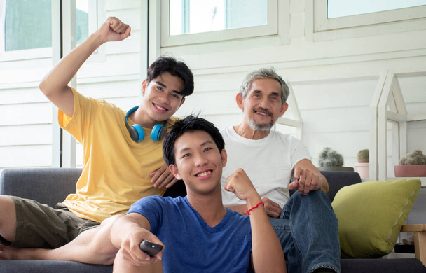 Grandsons and grandfather, sitting on sofa, holding remote tv, watching live soccer competition on tv programme and raise a fist up, concept for happy life of people in special times around the world. - Photo, Image