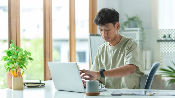 Νεαρός επιχειρηματίας κοιτάζοντας έξυπνο ρολόι χρόνο ελέγχου, ενώ κάθεται στο γραφείο στο σπίτι με φορητό υπολογιστή, το σχεδιασμό της εργασίας, έννοια διαχείρισης εργασιών. - Φωτογραφία, εικόνα
