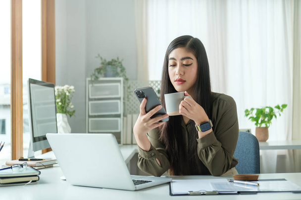 Okouzlující podnikatelka drží šálek kávy a pomocí mobilního telefonu v jasné moderní kanceláři. - Fotografie, Obrázek