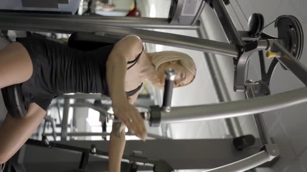 Mujer atlética hermosa y fuerte que utiliza simulador para levantar y extender las manos para bombear grupo de músculos en el gimnasio - Imágenes, Vídeo