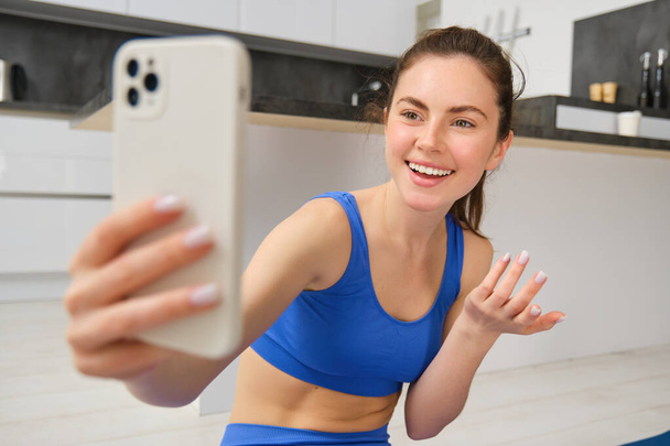Retrato de menina fitness jovem, blogueiro registra seu treino em casa, posa para selfie no smartphone, faz ioga na câmera, senta-se em esportista azul e leggings. - Foto, Imagem