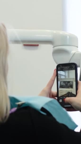 Blonďatá pacientka si vezme rentgen zubů na smartphone, zatímco sedí v zubním křesle. Mladý pacient v zubní ordinaci. Svislé video - Záběry, video