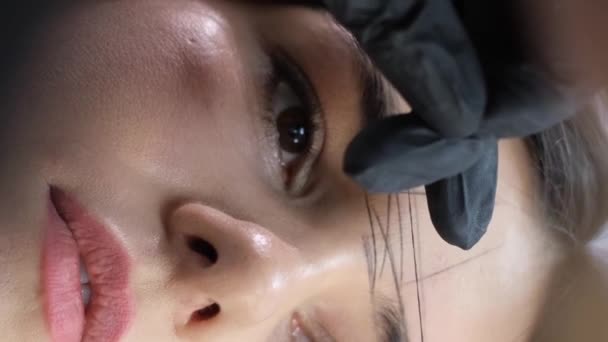 El cosmetólogo maestro corrige la forma de las cejas. Hermosa cara de una joven modelo en un salón de belleza antes de microblading, maquillaje permanente del tatuaje de la ceja. Vídeo vertical - Metraje, vídeo