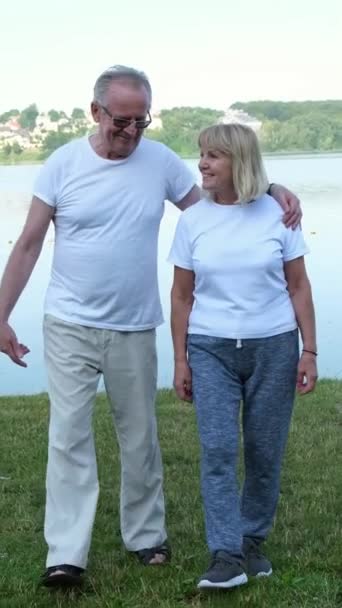 Aktiivinen onnellinen kypsä pari urheiluvaatteissa puhuu aamuharjoituksen jälkeen, lämpenee, seisoo lähellä järveä. Pystysuora video - Materiaali, video