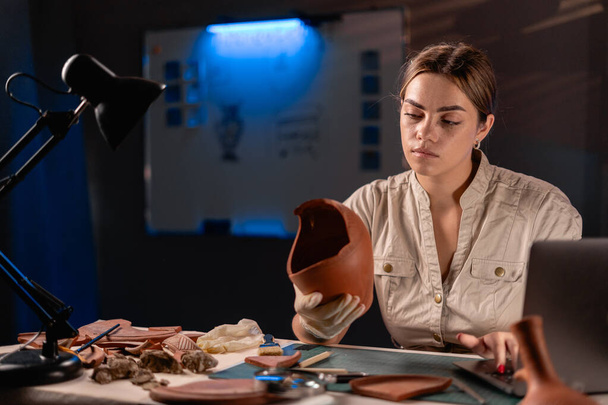 Γυναίκα αρχαιολόγος κάνει έρευνα που εργάζεται αργά το βράδυ στο γραφείο, χρησιμοποιώντας φορητό υπολογιστή, αναλύοντας αρχαία πολιτιστικά αντικείμενα πολιτισμού. Αντιγραφή χώρου - Φωτογραφία, εικόνα