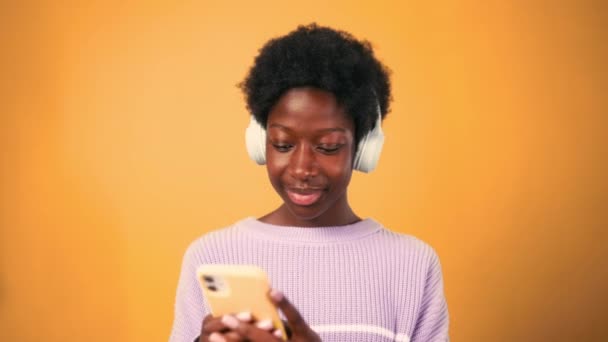 afro-américaine jeune femme avec afro coiffure debout en pull sur un fond orange vif portant écouteurs écouter de la musique et en utilisant le téléphone. - Séquence, vidéo