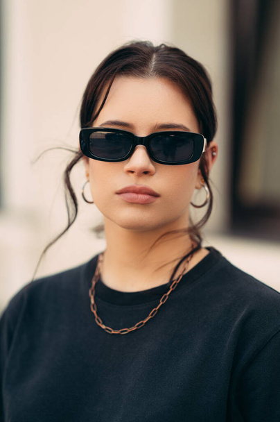 Függőleges szabadtéri portré egy komoly fiatal nőről fekete pólóban és napszemüvegben.. - Fotó, kép