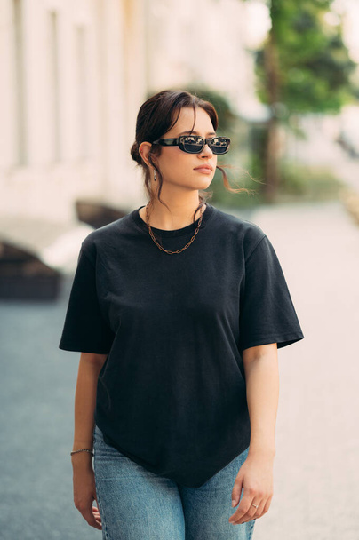 Verticaal buitenportret van een serieuze jonge vrouw die op straat loopt met een zonnebril op. - Foto, afbeelding