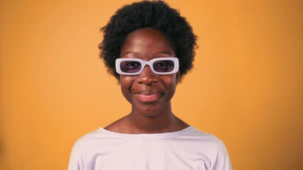 African American νεαρή γυναίκα με αφρο στυλ μαλλιά σε γυαλιά στέκεται σε πουλόβερ σε ένα φωτεινό πορτοκαλί φόντο και χαμογελώντας. - Πλάνα, βίντεο
