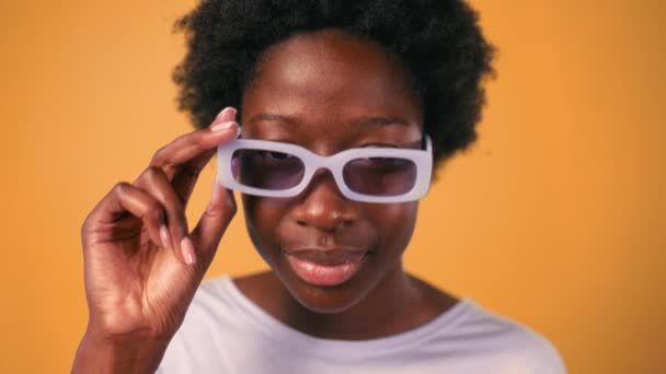 Una joven afroamericana con un peinado afro y gafas se coloca en un suéter sobre un fondo naranja brillante mirando quién está allí mientras se quita las gafas. - Imágenes, Vídeo