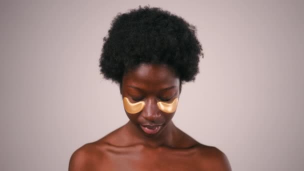 Mujer joven afroamericana con pelo afro y perfecta incluso la piel sana usando parches para los ojos aislados sobre fondo gris. Tratamientos de cuidado de la piel y spa. - Imágenes, Vídeo