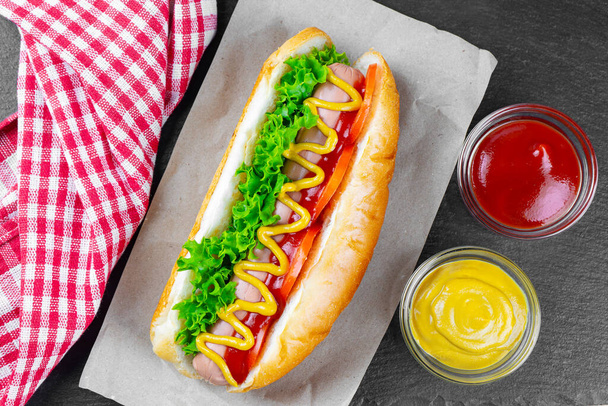 Hot Dog fatto in casa con senape gialla, ketchup, pomodoro e foglie di insalata fresca su sfondo di ardesia nera. Fast food, street food, cucina americana - Foto, immagini