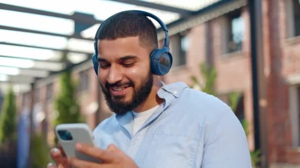 Close up of excited bearded man dancing and listening to music with wireless headphones on street. Content young guy in casual outfit holding personal smartphone in hands and scrolling list of tracks. - Footage, Video