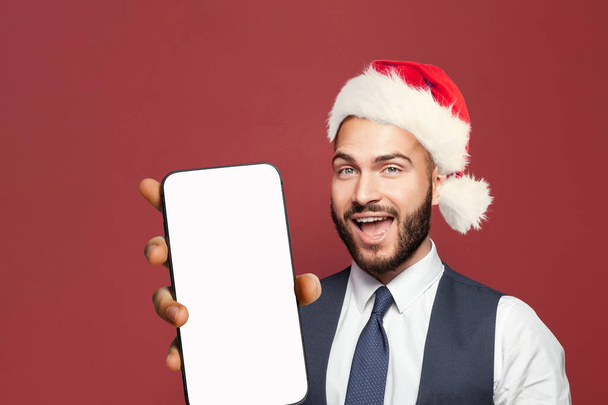 Рождественский бизнесмен в шляпе Санта Клауса показывает смартфон с пустым пустым экраном. Гай Санта играет в игры, серфинг онлайн или обучение. Экранный макет телефона Xmas - Фото, изображение