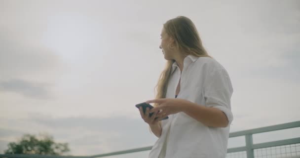 Αργή κίνηση πλάνο ελκυστική νεαρή γυναίκα που χρησιμοποιεί το κινητό τηλέφωνο ενάντια στον ουρανό - Πλάνα, βίντεο