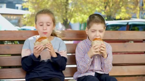 Malé děti, chlapec a dívka jedí hamburgery, pouliční jídlo sedí v létě na lavičce ve městě - Záběry, video