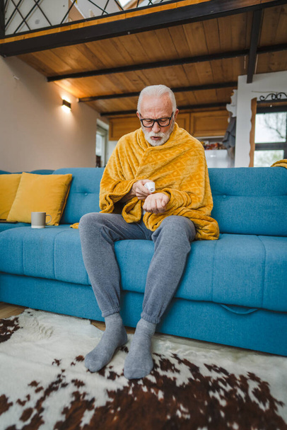 jeden biały mężczyzna starszy mężczyzna siedzieć na kanapie w domu trzymać termometr sprawdzić temperaturę ciała gorączka lub przeziębienie kichanie chore złapany zimno - Zdjęcie, obraz