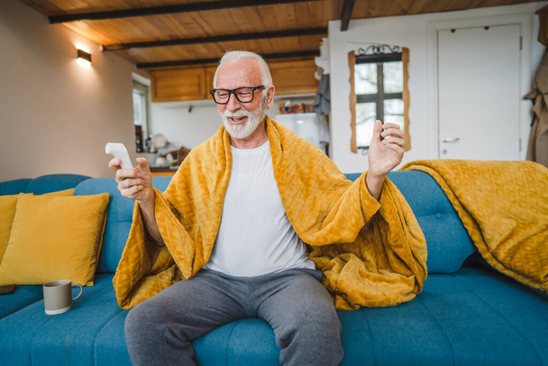ein kaukasischer männlicher Senior sitzt zu Hause auf dem Schlafsofa und hält Thermometer in der Hand, um die Körpertemperatur zu überprüfen, Fieber oder Schnupfen - Foto, Bild