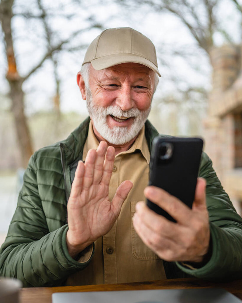 Yksi mies vanhempi valkoihoinen mies harmaa parta ja korkki istua ulkona keväällä tai syksyllä päivä onnellinen hymy käyttää mobiili älypuhelin videopuhelu kopioi tilaa oikeita ihmisiä - Valokuva, kuva